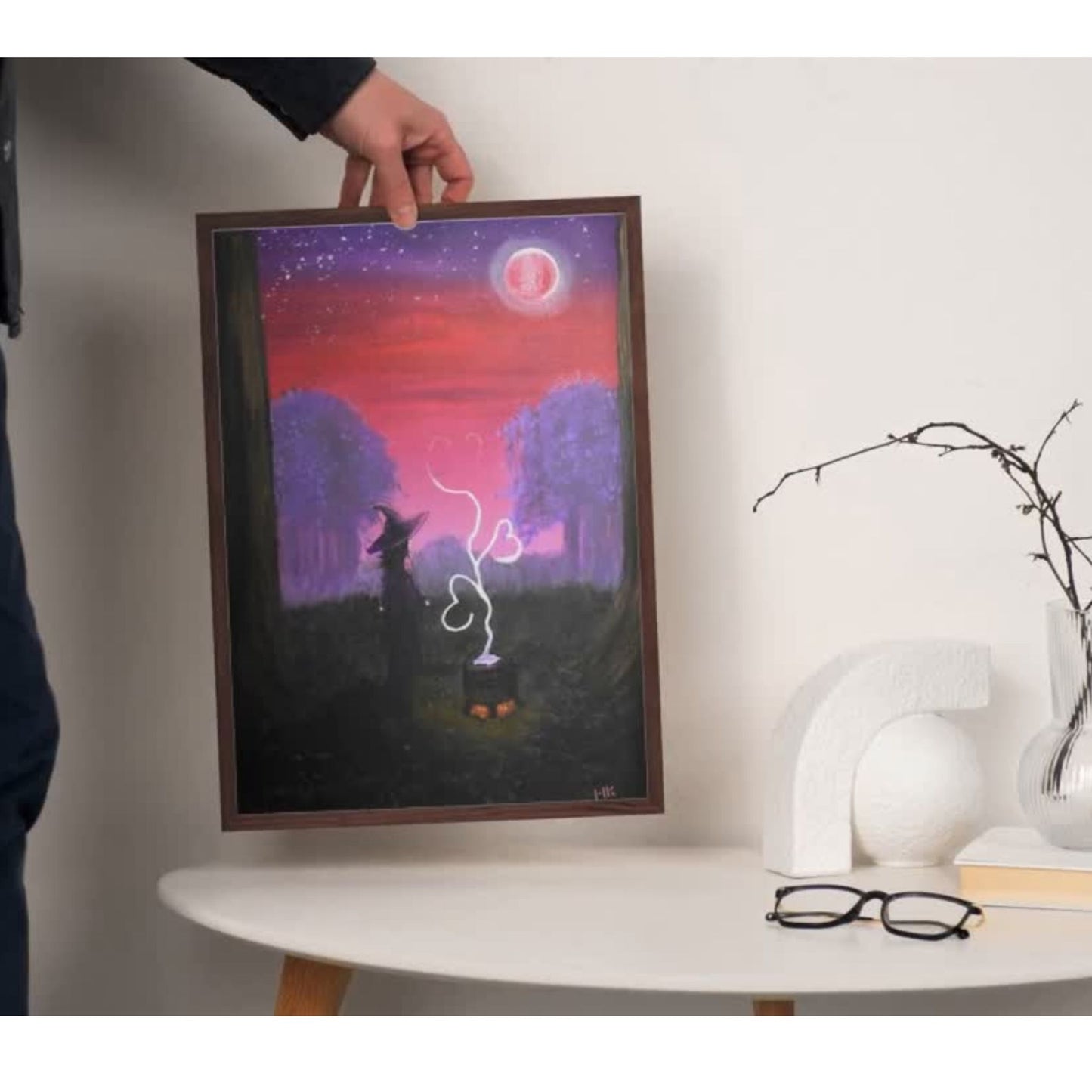 Framed artwork of a witch and cat silhouette against a purple sky with a red moon, held by a person.