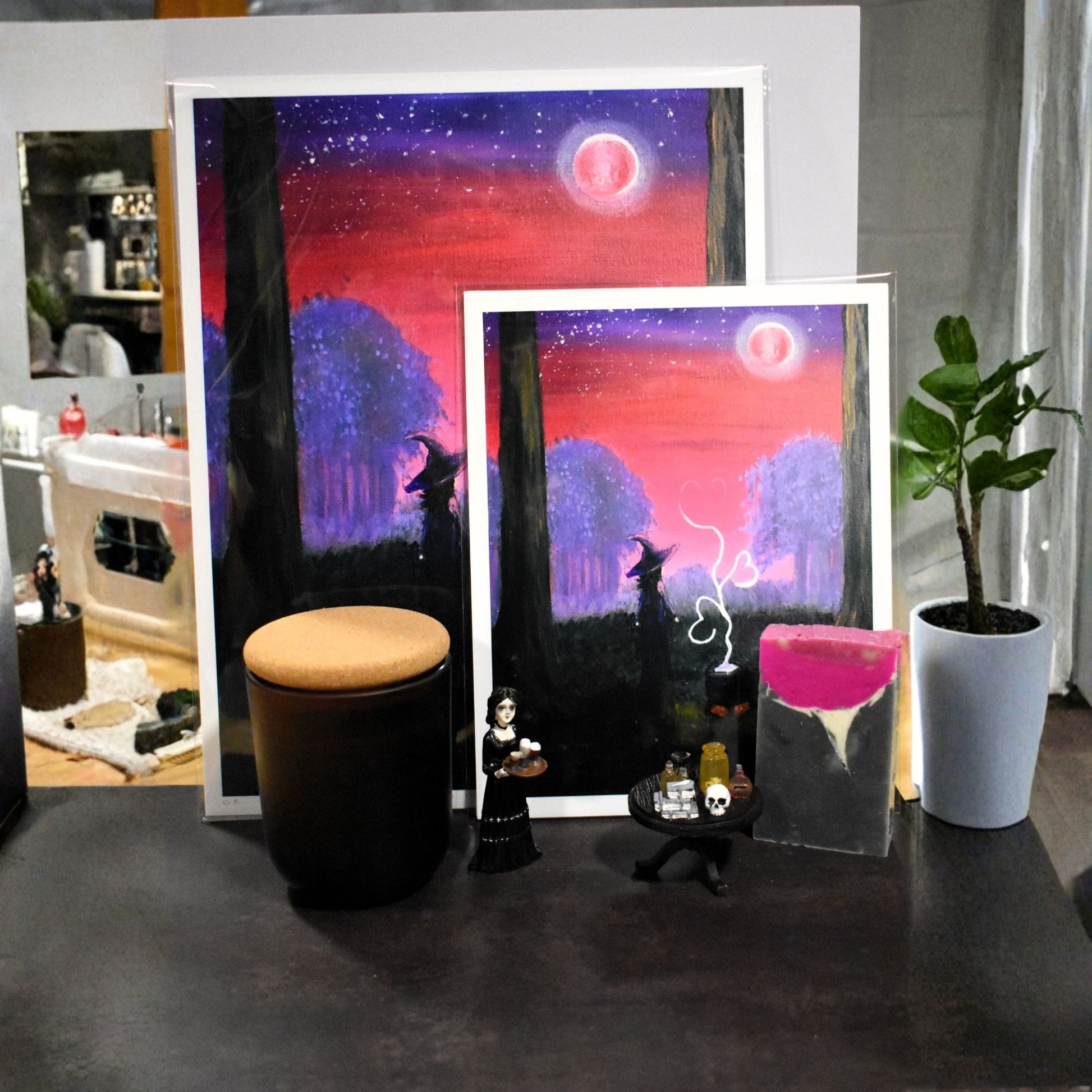 Two framed artworks on a table with a plant and small stool in the background.